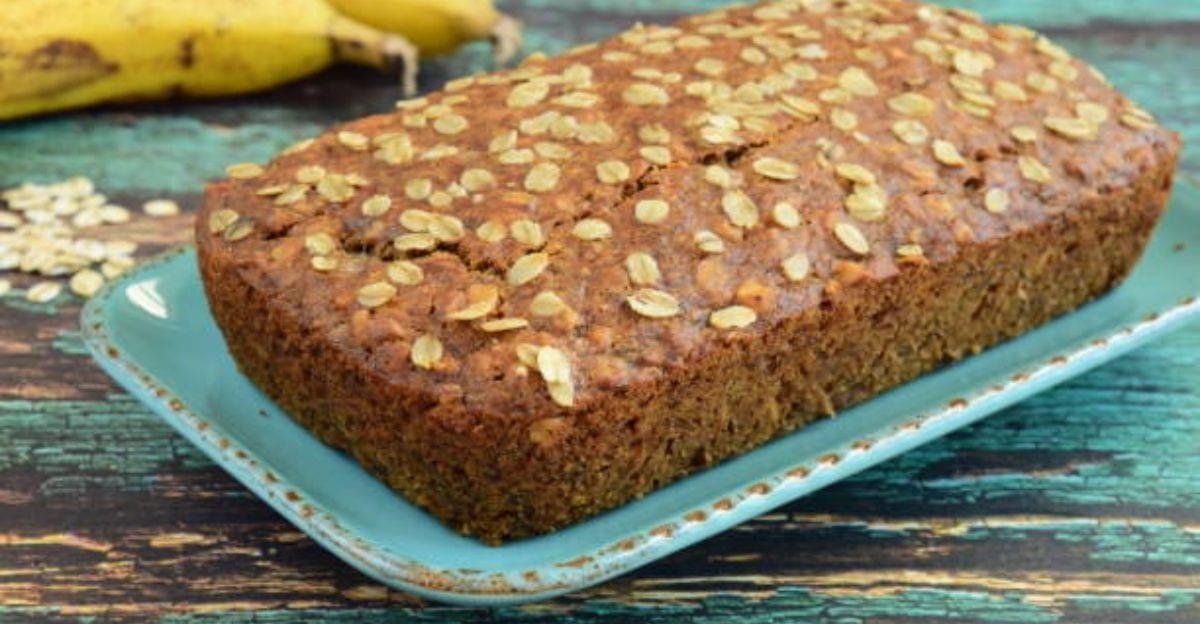 Freshly baked banana oat cake sliced on a wooden board with ripe bananas and rolled oats beside it.